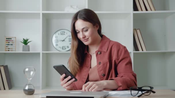 Donna affascinante con smartphone leggere messaggi dal fidanzato sorridente in seduta in ufficio. Mozione. Persone urbane. — Video Stock