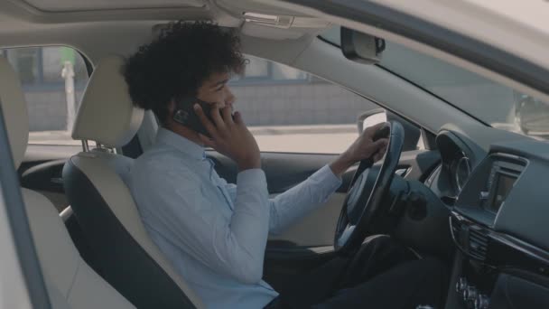 सुंदर युवा अफ्रीकी अमेरिकी आदमी सामने के चमड़े की सीट पर बैठे काले लक्जरी कार और अपने मोबाइल पर बात करते समय वाहन टच स्क्रीन ट्यून करता है — स्टॉक वीडियो