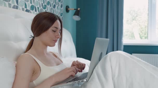 Mujer morena trabajando ordenador portátil en la cama. Feliz bonita mujer trabajando desde casa en el ordenador portátil clave desde el dormitorio. — Vídeos de Stock