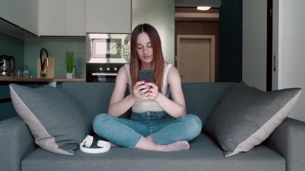 リラックスしたブルネットの若い女性は、ソーシャルメディアをサーフィンするスマートフォンを使用して、ニュースをチェックし、モバイルゲームや居心地の良いソファに座っているテキストメッセージを再生します。千年の女性は細胞と一緒に家で時間を過ごす — ストック動画