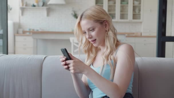 Ontspannen blonde jonge vrouw met behulp van smart phone surfen sociale media, het controleren van nieuws, het spelen van mobiele games of sms-berichten zitten op gezellige bank. Millennial dame besteedt tijd thuis met mobiele gadget — Stockvideo