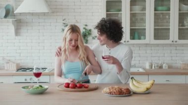 Mutlu ve genç bir çift taze sebze salatası kesiyor. Mutfakta birlikte yemek pişiriyor. Gülümseyen koca, kırmızı şarap içiyor ve karı koca olarak gülüyor.