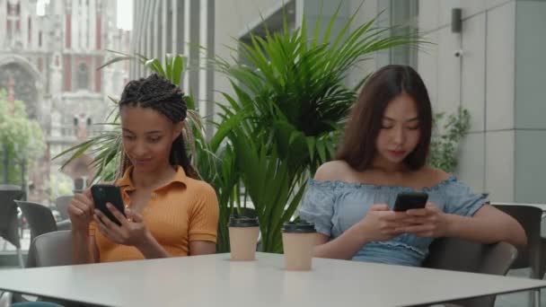 Asiatiques et mixtes ra e jeunes filles tenant smartphone utilisent les médias sociaux boire du café dans un café tout en étant assis au café à l'extérieur et pointant du doigt. Concept de dépendance à Internet — Video