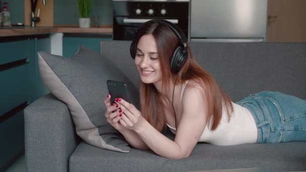Donna attraente ascoltare musica indossando cuffie rilassarsi sul divano sms utilizzando smartphone navigazione online giocare giochi per cellulare godendo di condividere messaggi sui social media 4k — Video Stock