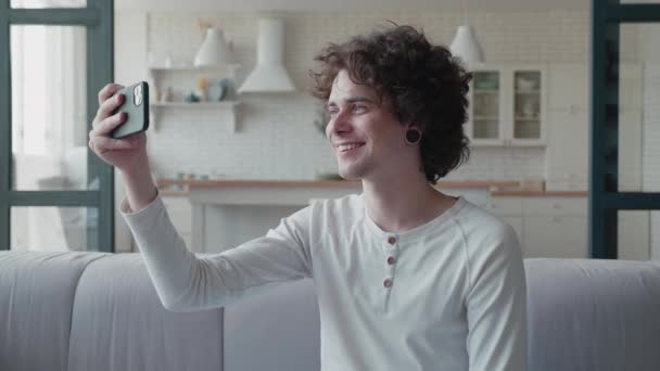 Χαρούμενος χιλιετής τύπος με σήραγγες αυτιών και σκουλαρίκια στη μύτη συνομιλεί με βιντεοκλήση με φίλους και χαμογελώντας κοιτάζοντας κάμερα του smartphone, ή blogging κάθεται μόνος στο σπίτι, συνομιλία βίντεο — Αρχείο Βίντεο