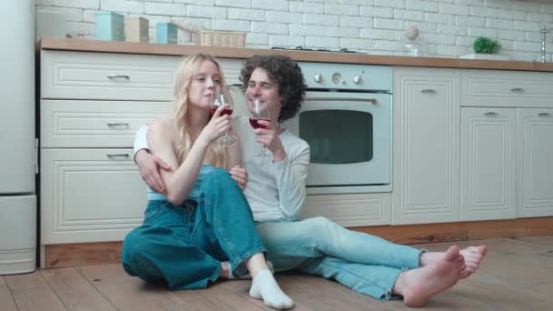 Pareja cariñosa feliz joven y mujer rubia sentarse en la acogedora sala de cocina piso celebrar copas beber vino tinto hablar risa celebrar vacaciones nueva casa comprar alquiler hipoteca inversión — Vídeo de stock