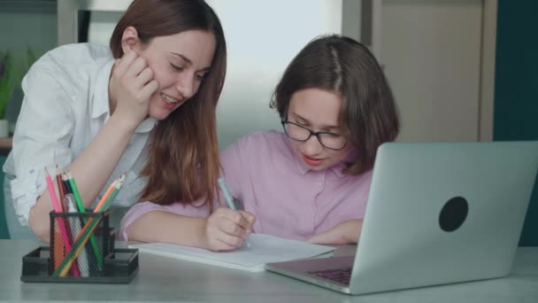 Vážná dcera dělá domácí úkoly vedle matky u stolu. Mladá máma pomáhá dítěti s úkoly doma. Dospívající dívka v brýlích studium dálkové s podporou rodičů uvnitř — Stock video