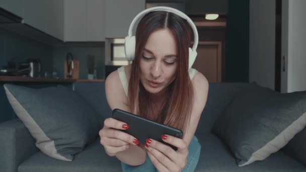 Jonge vrouw speelt video game in smartphone via internet, met behulp van een koptelefoon en zitten op de bank in de vakantie tijd. Winnend concept — Stockvideo