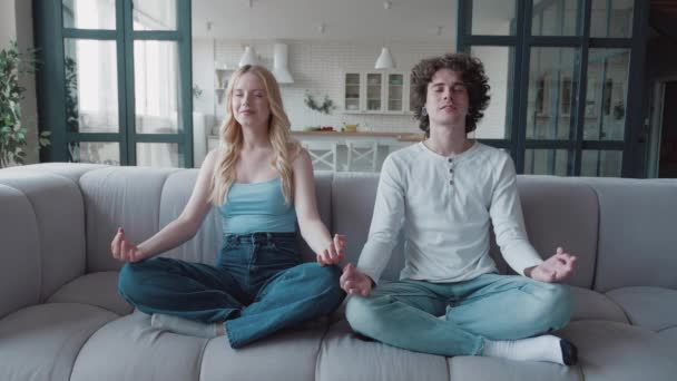 Glada par gör morgonträning hemma moderna loft vardagsrum på soffan. Kaukasiska unga kvinna och man gör samma rörelse med armarna sitter i Lotus utgör meditater, andliga övningar lektioner — Stockvideo