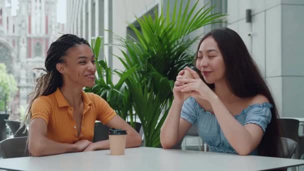 Joven ébano mezclado chica de raza con rastas y atractivo asiático chica sentarse en una mesa y tener lindo conversación al aire libre. Dos mujeres jóvenes hablando y tomando café sentadas en la cafetería. — Vídeo de stock