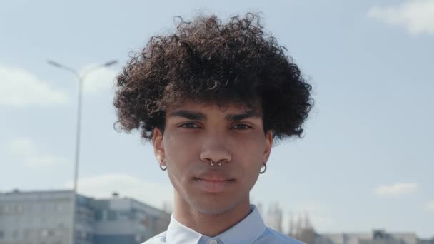 Primer plano joven divertido hombre africano pelo rizado de 20 años de edad lleva camisa blanca azul posando mirando cámara encantadora sonrisa de pie en la calle. Gente emociones sinceras concepto de estilo de vida — Vídeo de stock