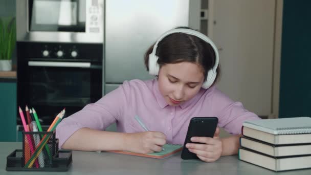 Cute child girl wearing headphones studying online in mobile app by video call with remote teacher, tutor. School kid holding phone doing homework using application, watching class learning at home. — Stock Video