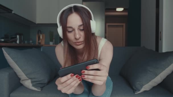 Y-generációs nő fejhallgatóval ül egy okostelefonon. Nő online játék okostelefon interneten — Stock videók