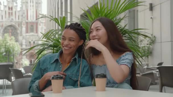 Des amies noires et asiatiques buvant du café ensemble, parlant et souriant. Jeune femme assise dans un café d'été et commérages en plein air — Video