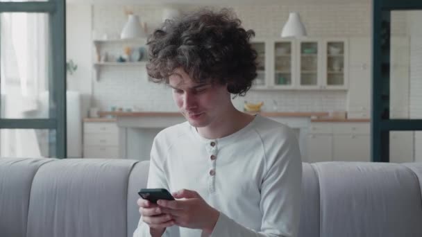 Gelukkig ontspannen jonge man met smart phone kijken naar mobiele telefoon scherm lachen genieten van het gebruik van mobiele apps voor winkelen plezier spelen van games chatten in sociale media zitten op gezellige bank thuis — Stockvideo