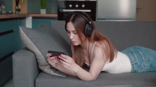 若い女性はインターネットでスマートフォンでビデオゲームをしていますヘッドフォンを使用して休暇時にソファの上に横たわっています。勝利の概念 — ストック動画
