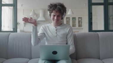 Kulaklıklı, gülümseyen ve pozitif hipster bir adam modern dairelerde bilgisayarla video görüşmesi yapıyor. Erkek çalışan web kamerasına el sallıyor. Video konferansı yapıyor.
