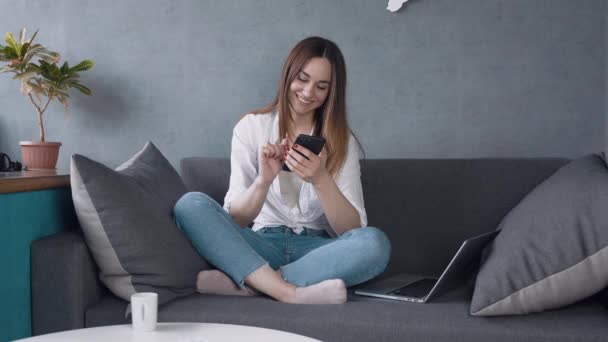 Молода жінка використовує смартфон серфінг соціальних мереж, перевіряє новини, грає в мобільні ігри або смс-повідомлення, сидячи на дивані . — стокове відео