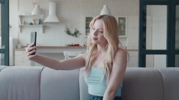 Ung blond kvinna gör selfie på sin smartphone sitter i mysig soffa i modern lägenhet — Stockvideo