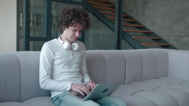 Ung kille i lutar sig mot bekväm soffa, arbetar på distans på datorn hemifrån. Fokuserad tusenårig student studerar distans på online-kurser. — Stockvideo