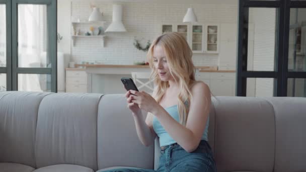 Mujer rubia feliz usando teléfono inteligente sentado en el sofá en la acogedora sala de estar elegante, chat y mensajería en línea, pasar el ocio en las redes sociales, compartir noticias con amigos — Vídeos de Stock