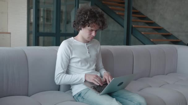 Geconcentreerde krullende man typt op laptop terwijl hij op de bank zit in het modieuze appartement. Afstandsonderwijs of freelance concept — Stockvideo