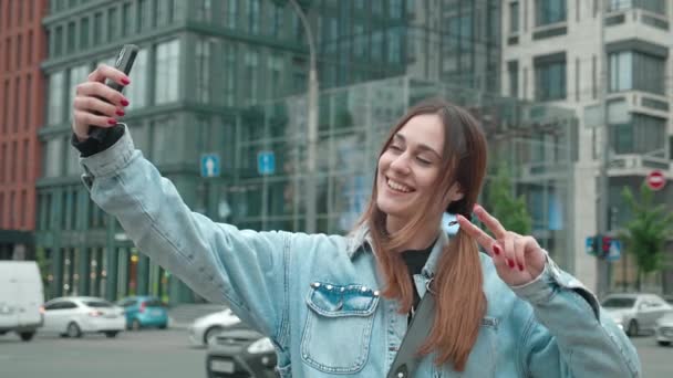 Junge Frau in Jeansjacke macht Selfie vor städtischem Hintergrund. Lustige Frau lächelt in Kamera Smartphone und zeigt Frieden — Stockvideo