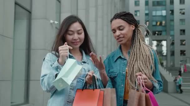 Twee gelukkige jonge Aziatische en Afrikaanse vrouwen wandelen door de stad na het winkelen op een straat met cadeauzakjes in koud of lenteweer 2 vrolijke meisjes gaan naar buiten van een uitverkoop — Stockvideo