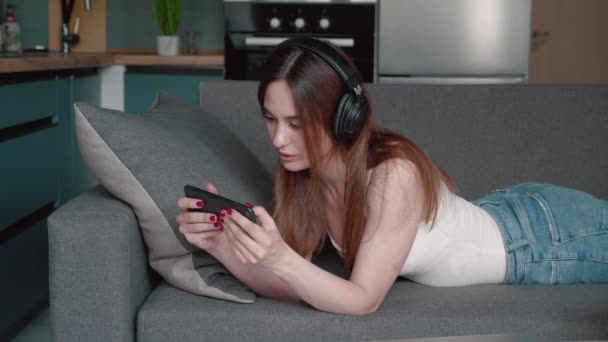 Lustige Frau spielt Videospiel im Smartphone per Internet, benutzt Kopfhörer und liegt zur Urlaubszeit auf der Couch. Win-Konzept — Stockvideo