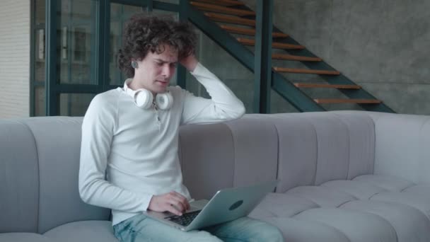 Relaxado sério millennial cara estudante freelancer usando dispositivo laptop inclinado no sofá em casa escritório, empreendedor focado trabalhando distantemente digitando no notebook no apartamento — Vídeo de Stock