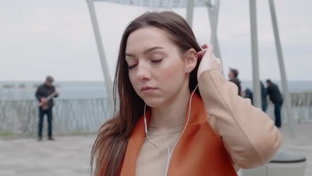 Een aantrekkelijke vrouw zet een oortelefoon op en luistert naar muziek op het buitenterras. Ze drinkt koffie. vrijetijdsconcept — Stockvideo