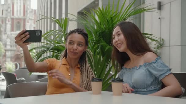 写真を撮るか、街の背景にあるカフェに座っているスマートフォンの友人とビデオチャットをしている2人の幸せなガールフレンド — ストック動画