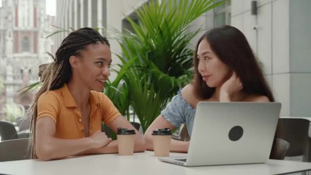 Dos chicas jóvenes de raza mixta tienen una linda conversación mientras están sentadas al aire libre en un café de verano del fondo urbano de la ciudad — Vídeo de stock