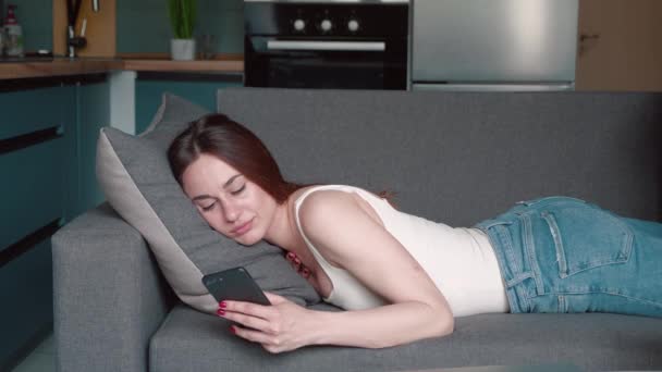 自宅の服でリラックスした若いブルネットは手にスマートフォンを持つ灰色のソファに横たわっています。魅力的な女の子リラックスして自宅で現代的なガジェット. — ストック動画