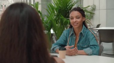 Güneşli bir günde dışarıda oturmuş kahve içerken sohbet eden iki iş arkadaşı. Kot ceketli Afrikalı Amerikalı kadına odaklan.