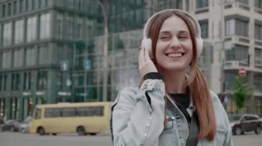 Kulaklık takmış genç bir esmer kadın müzik dinliyor ve şehrin arka planında eğlenceli bir şekilde dans ediyor. Komik kadın yalnız eğleniyor, mutlu ve kaygısız hissediyor. Neşeli dans. Eğlence ve