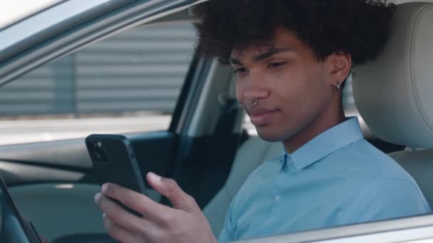 Estudiante afroamericano está utilizando el teléfono inteligente, sentado en el coche, la comunicación en las redes sociales, usuario masculino de gadget. Joven chico escribiendo mensaje — Vídeos de Stock