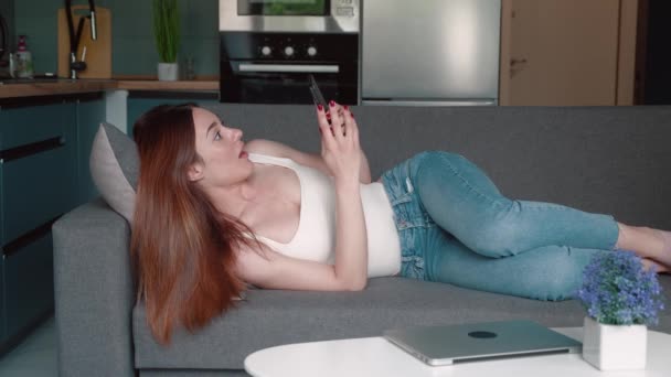Chica tumbada relajándose en el sofá y desplazándose con su teléfono inteligente y recibirá mensajes largamente esperados — Vídeo de stock