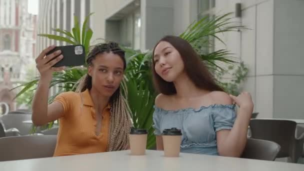 二つの幸せなガールフレンドは、友人とのビデオチャットを持っている都市の背景にあるカフェに座っているスマートフォンのカメラであなたの手を振る — ストック動画