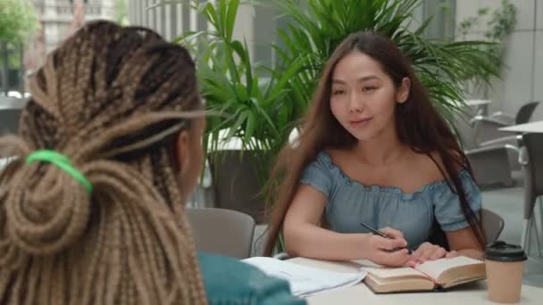 Focus jeune femme asiatique avec livre ses notes.Two collègues ou amis assis à l'extérieur le jour ensoleillé, parler autour du café — Video