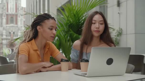 Laptoplu iki güzel kız şehrin arka planındaki kafede oturuyor. Karışık ırk kızı ve Asyalı kadın. — Stok video