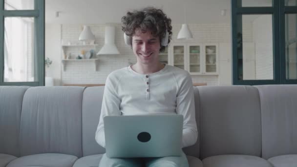 Привлекательный кудрявый парень сидит на диване в светлой комнате дома с ноутбуком счастливы говорить по видеосвязи. Женщина общается в Интернете и улыбается, активно обсуждает новости с другом — стоковое видео