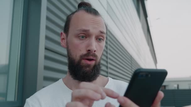 Homme surpris regardant dans le smartphone. Beau mec avec une expression wow. Gros plan jeune homme posant sur le fond. Portrait d'un étudiant souriant près du bâtiment — Video