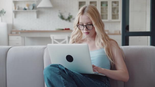 Bela estudante loira com óculos e jeans está estudando remotamente em uma grande sala brilhante em casa com um laptop no colo. A menina sentada sofá aconchegante e digitando em um laptop — Vídeo de Stock