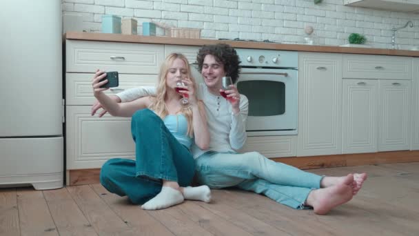 Kochająca para szczęśliwy młody facet i blondynka biorąc selfie siedzieć na przytulnej kuchni pokój piętro trzymać szklanki pić czerwone wino rozmawiać śmiech świętować wakacje nowy dom zakup wynajem hipoteki inwestycji — Wideo stockowe