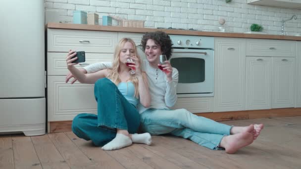 Pareja cariñosa feliz joven y mujer rubia tomando selfie sentarse en acogedora cocina habitación piso celebrar vasos beber vino tinto hablar reír celebrar vacaciones nueva casa comprar alquiler hipoteca inversión — Vídeo de stock