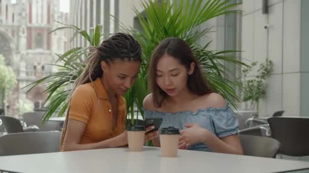 Deux jeunes filles utilisant un téléphone intelligent au café extérieur. Deux femmes après avoir fait du shopping assis dans un café en plein air avec du café et en utilisant un smartphone — Video