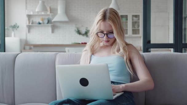 Una atractiva chica rubia con gafas sosteniendo el portátil en su regazo sentada en un sofá en un apartamento moderno — Vídeo de stock