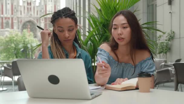 Die charismatische Studentin y und ihre Kollegin mit den langen Haaren sitzen nach dem College gemeinsam auf der Straße im Café. Afroamerikanische amerikanische und asiatische Mädchen suchen Laptop und schreiben in ein Notizbuch — Stockvideo