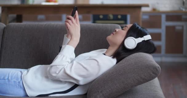Feliz estudiante atractiva chica asiática está escuchando música en los auriculares que sostienen la pantalla táctil del teléfono inteligente tumbado en el sofá en casa. Gadgets, concepto de juventud y ocio. — Vídeos de Stock